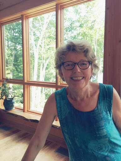 Hannelore, a seasoned Yoga Therapist and Ayurvedic practitioner, exuding warmth and serenity, embodies the essence of yoga and holistic wellness. In this image, she is seen wearing a blue sleeveless shirt, smiling at the camera, with a beautiful forest visible through large windows behind her. A plant on the window sill adds a touch of nature to the scene.