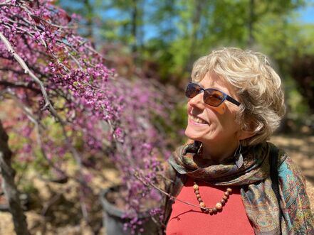 Smiling Hannelore standing in a beautiful forest, captivated by a branch of vibrant purple flowers on a tree. She wears a bright red scarf, adding a pop of color to the serene natural setting.